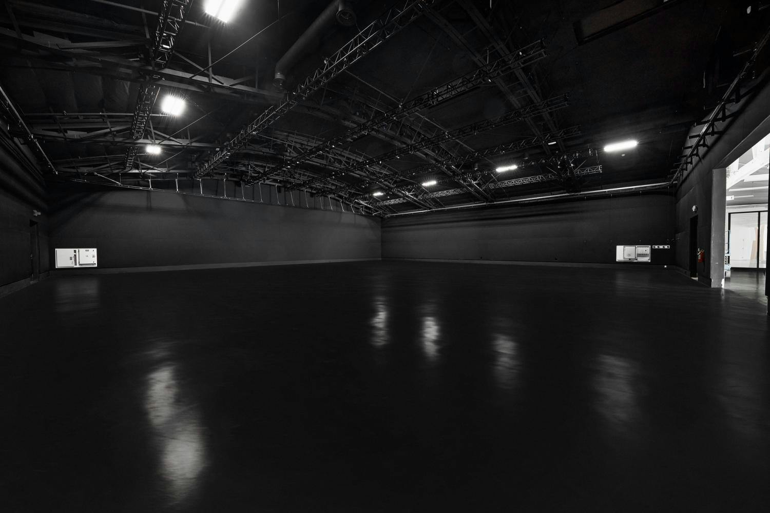 An expansive black-walled studio with high ceilings equipped with technical stage lighting and a reflective floor.