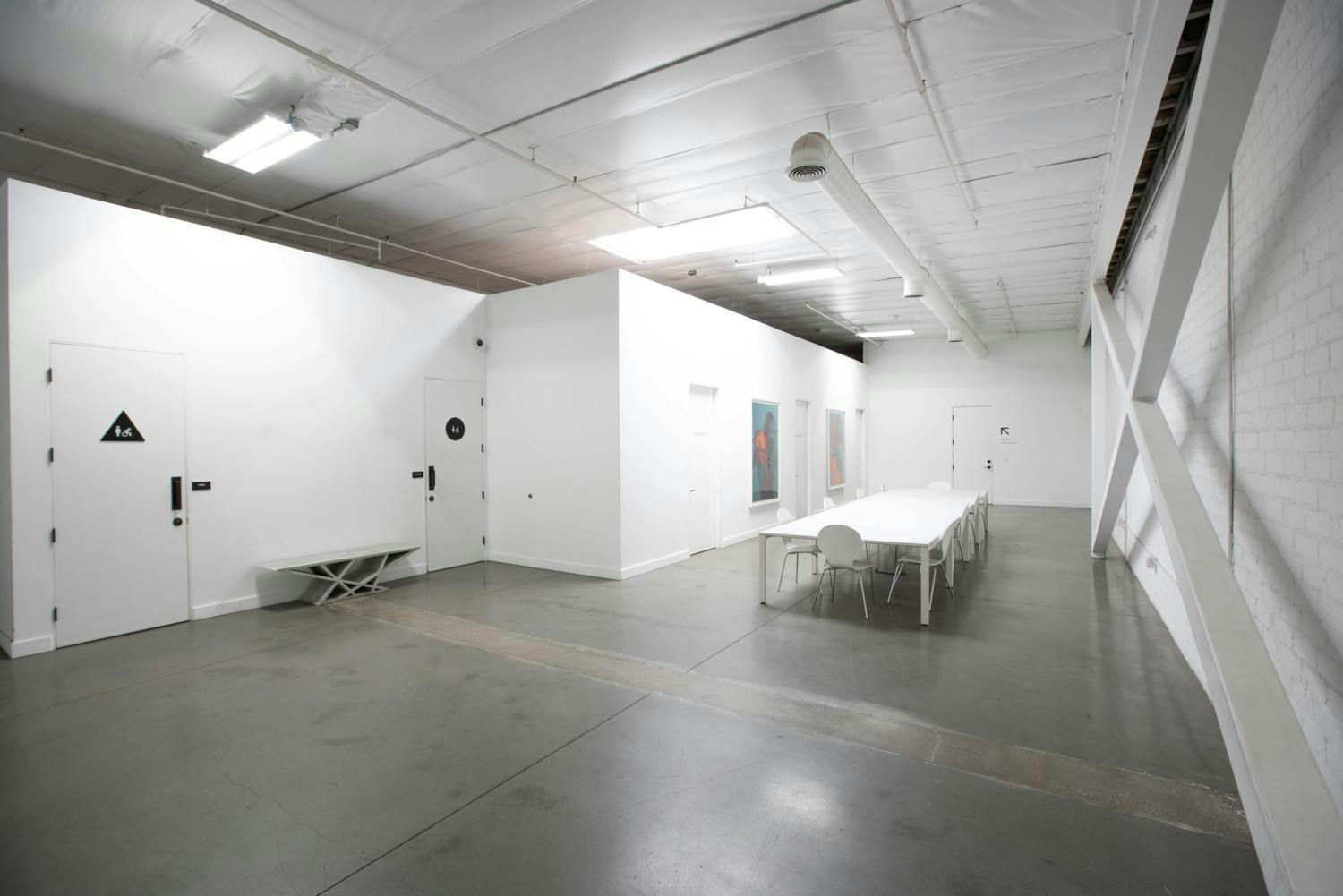 A bright and spacious gallery-like room with white walls, a long meeting table, and modern art, featuring an industrial ceiling with exposed pipes and ductwork.