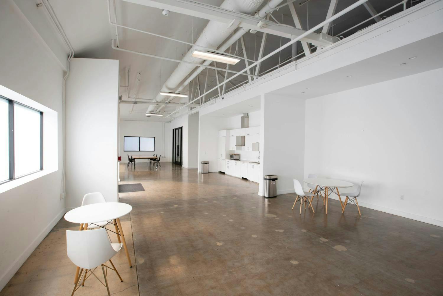 A clean and airy break area featuring a small round table with white chairs, a kitchenette, and a conference room in the background, in a modern studio space with high ceilings.