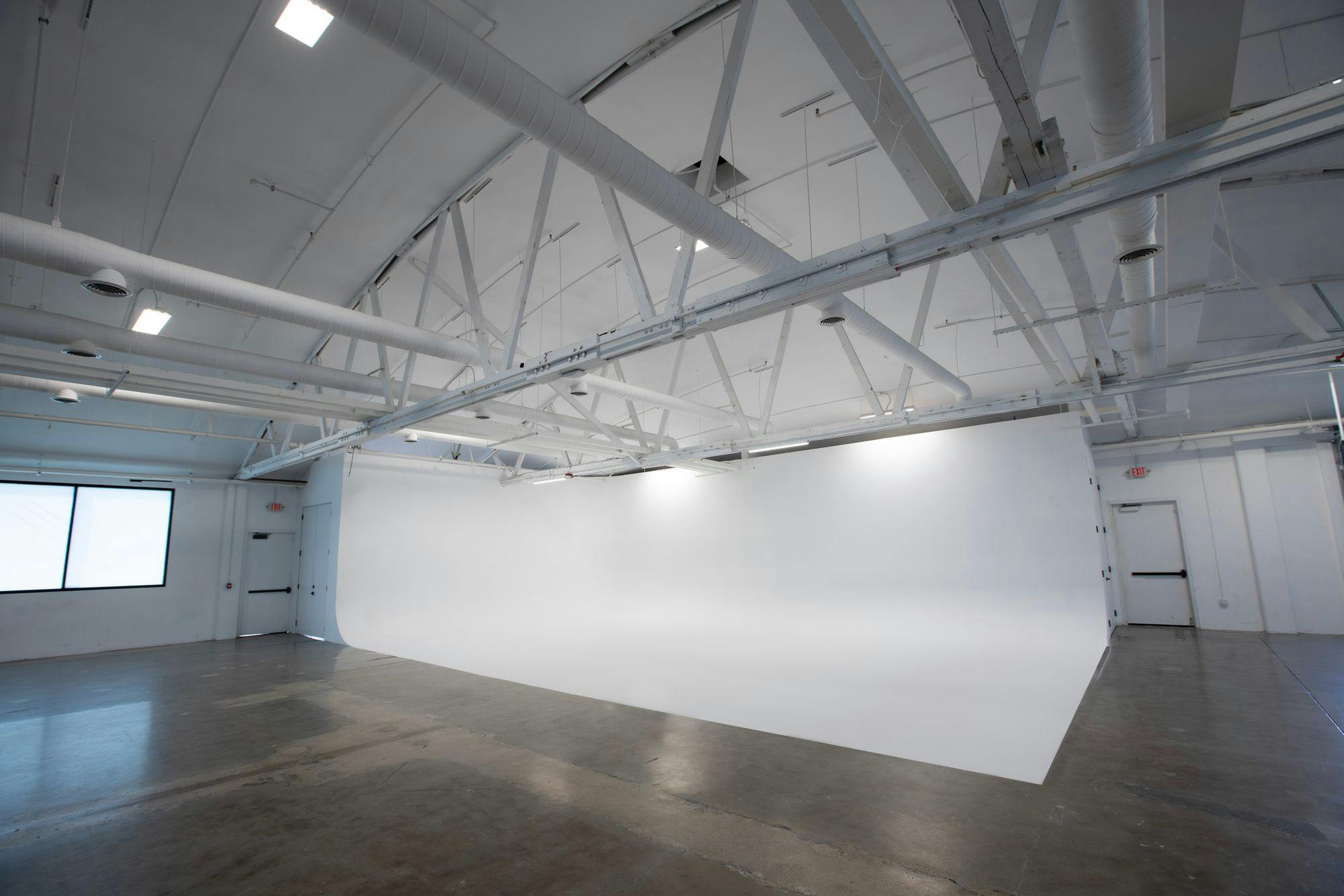A spacious industrial-style gallery with white walls and high ceilings, featuring a large blank white cyclorama on the right side and a concrete floor.