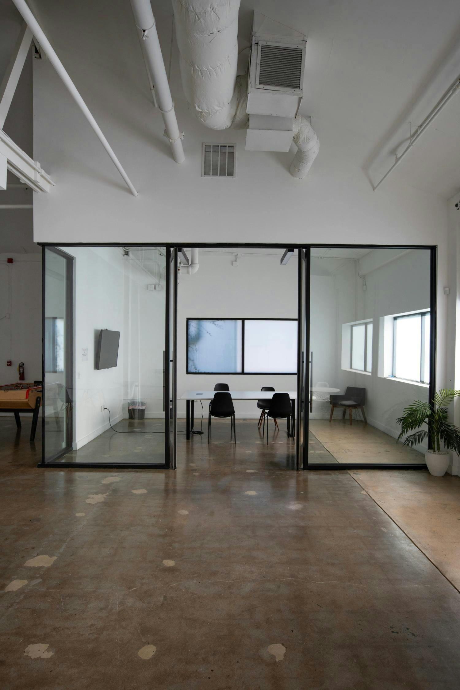 A shot of a modern studio space with glass partitions, a conference area in the view, and an urban industrial ceiling with exposed pipes and ductwork.