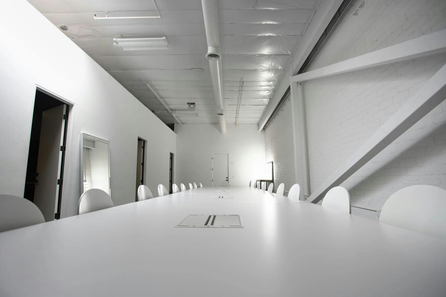 A modern meeting room with a long white table, white chairs, and white brick walls, complemented by industrial ceiling elements and ductwork.