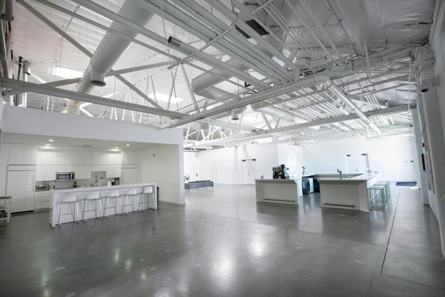 An expansive gallery with polished concrete floors, high ceilings with exposed ductwork, and a kitchen and seating area in the distance.