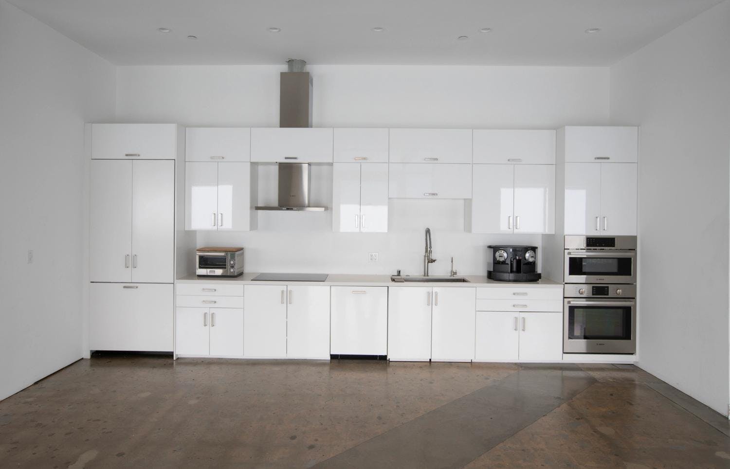 A modern studio kitchen featuring white cabinetry, stainless steel appliances, a microwave, an oven, and a coffee machine set against a white wall, with polished concrete floors.