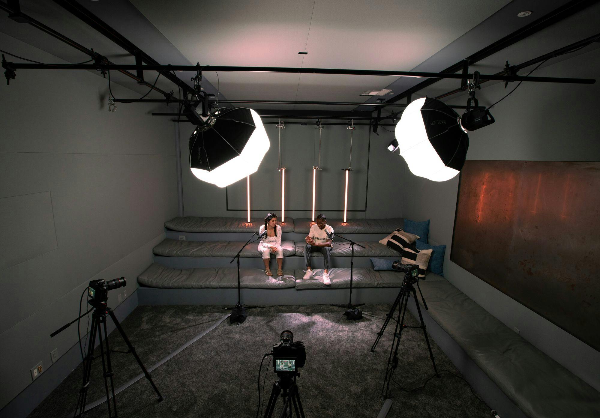 A podcast studio setup with two large softbox lights and a camera on a tripod, focusing on two people sitting on a couch against a backdrop with vertical tube lights.