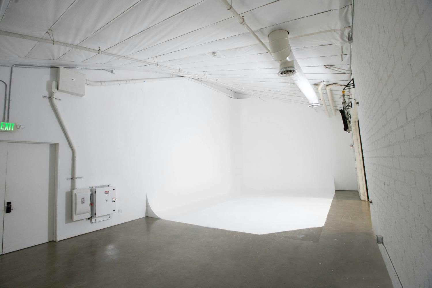 An empty photography studio with white walls and ceiling, featuring an HVAC duct, electrical panel, and concrete floor.