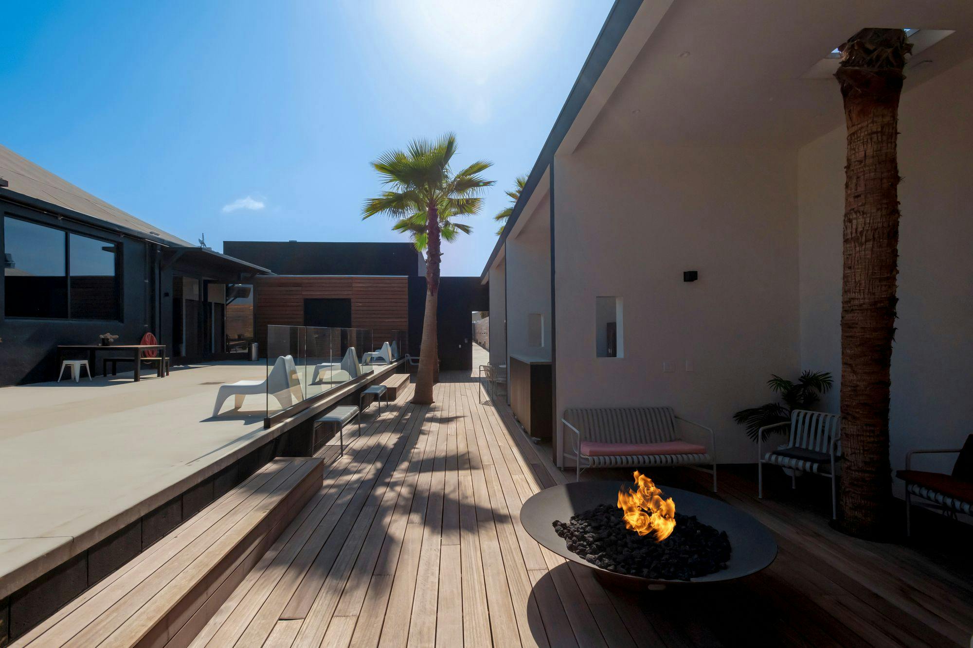 The photo displays an outdoor patio with a wooden deck, modern fire pit, sleek furniture, and a pair of palm trees, set against a backdrop of contemporary buildings.