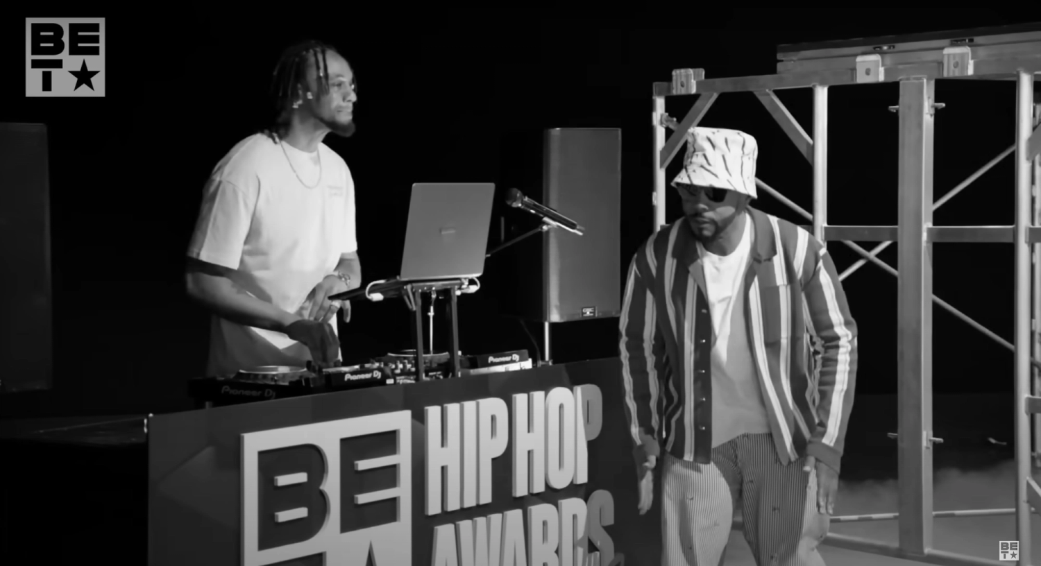 Two focused men, one DJing and the other poised to perform, at the "BE Hip Hop Awards," captured in a dramatic black and white photo.