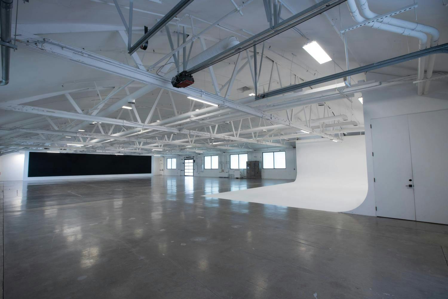 A corner view of a spacious, well-lit studio featuring a large black backdrop and a white cyclorama, with high ceilings and exposed white ductwork, ideal for a variety of media productions.