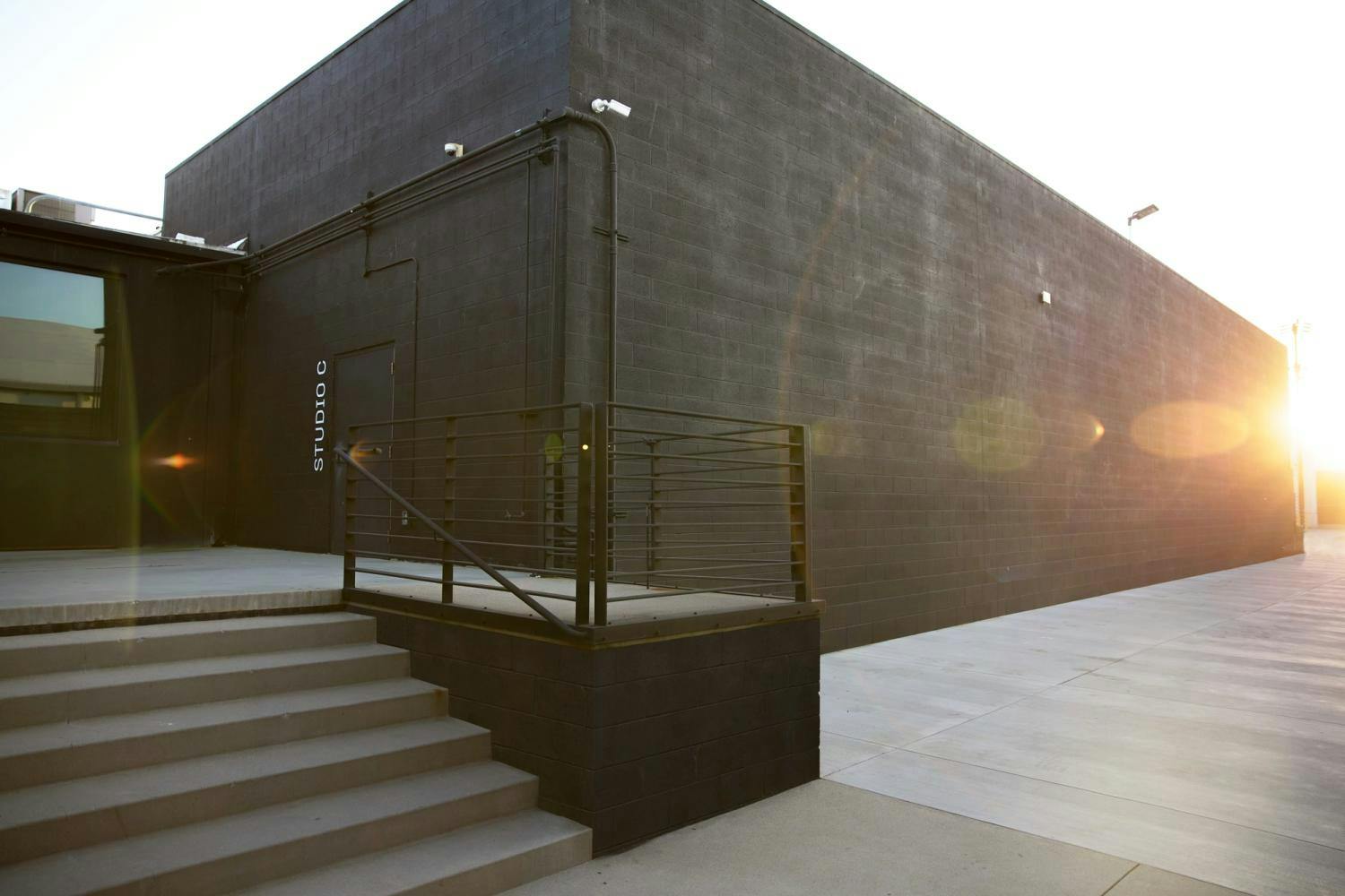 The exterior of Studio C captured at sunset, featuring concrete stairs, a railing, and a black brick wall, with the sun's glare adding a warm ambiance.