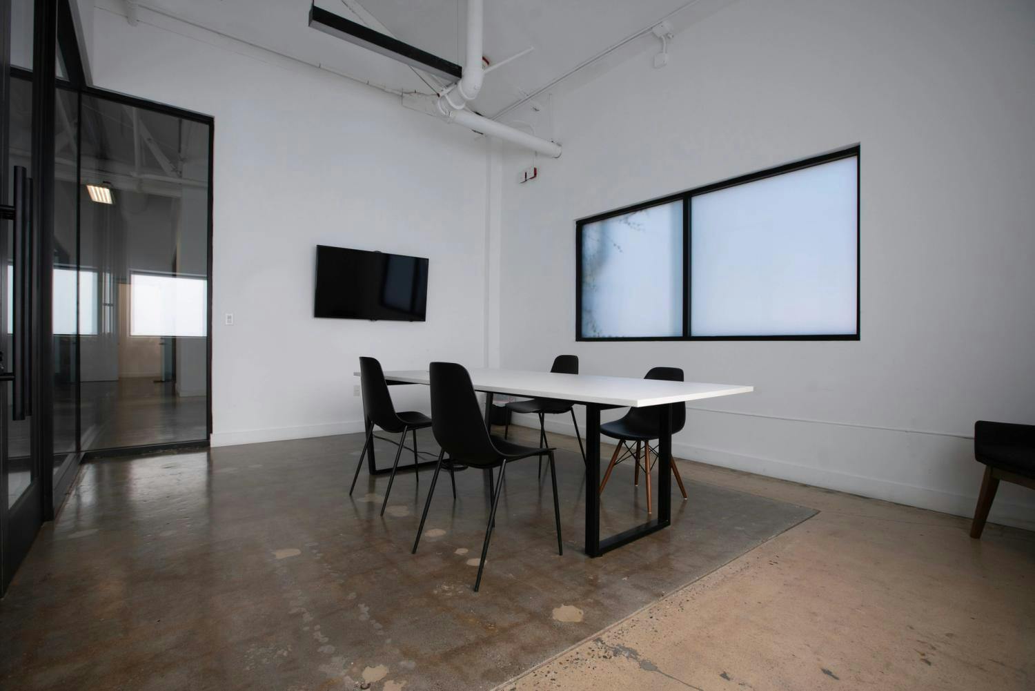 A modern meeting room with a white table, black chairs, and a wall-mounted TV, featuring an industrial-chic interior with glass doors and concrete floors.