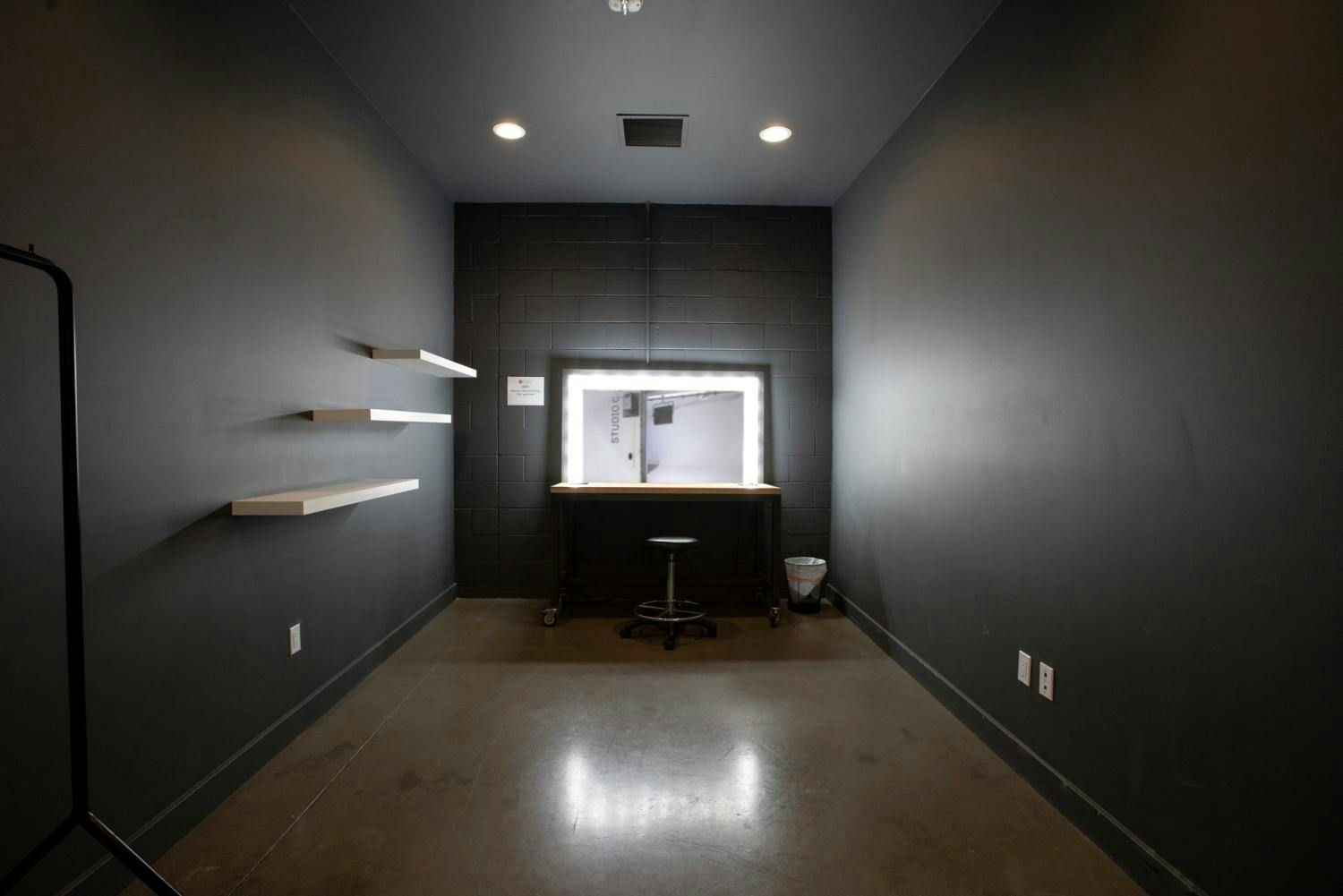 A dark-toned makeup room with a lit mirror, wooden desk, adjustable stool, and empty shelves on a gray wall.
