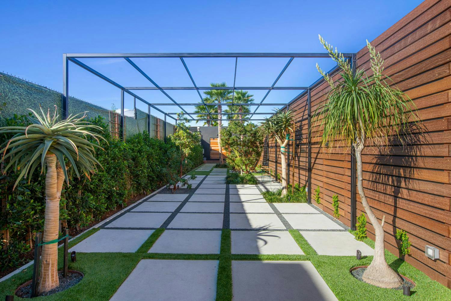 A landscaped garden path with symmetric stepping stones, flanked by wooden fences and exotic plants, under a metal pergola.