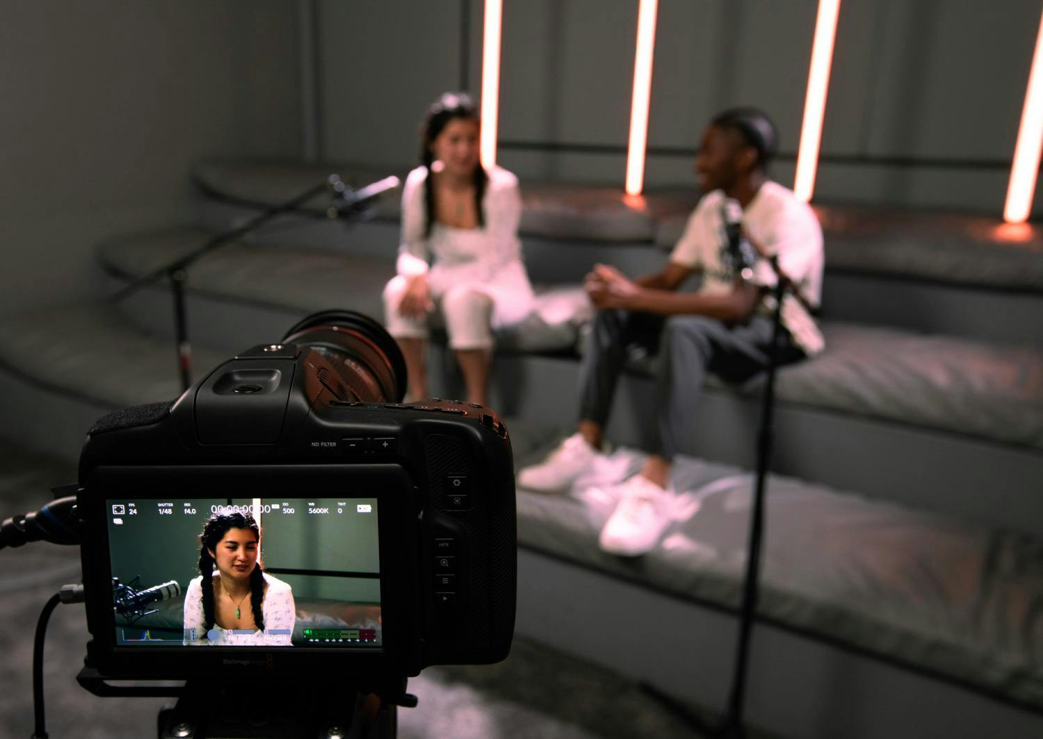 A camera focuses on a female interviewee in a modern studio with neon lights and a soft gray background.
