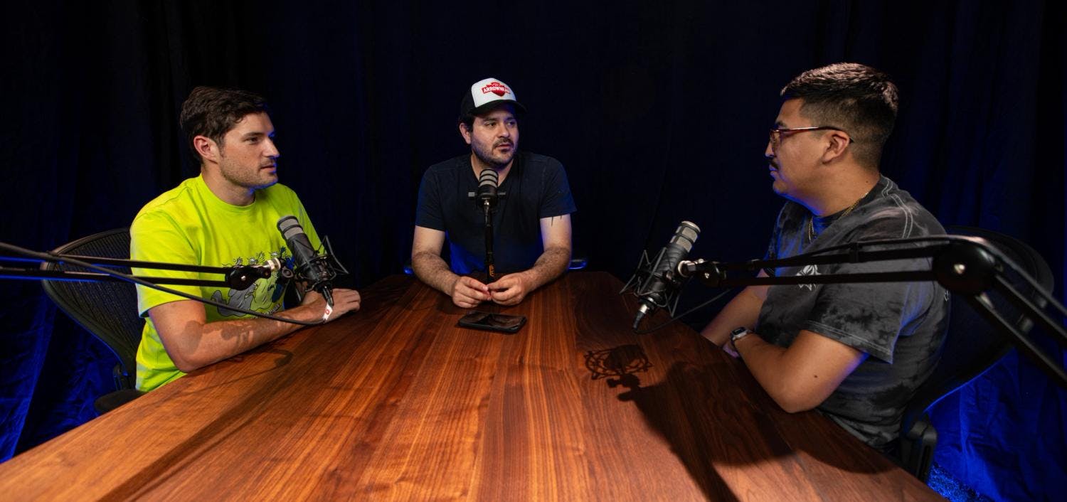 Three podcasters in a conversation at a round wooden table, with microphones and a black backdrop.