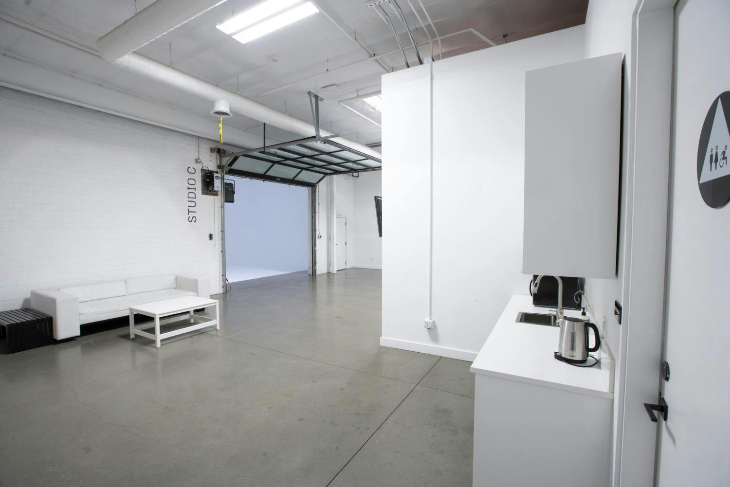 An airy studio break area with minimalist white furniture, a coffee station, and large open space leading to another room, marked by an overhead "STUDIO C" sign.