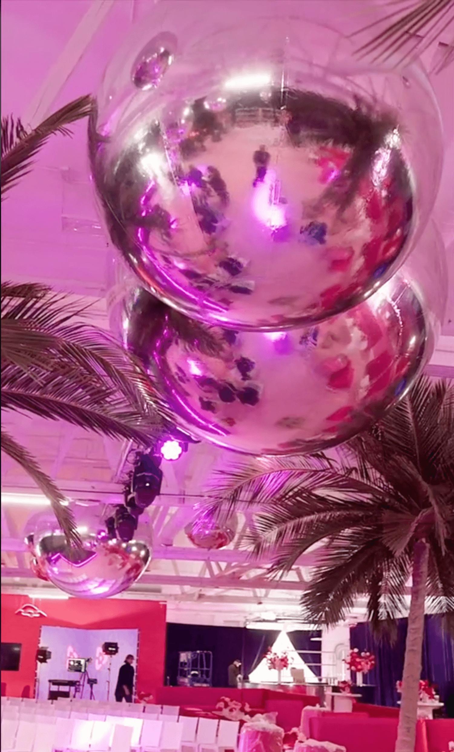 Giant reflective balloons hover above a lively event space decorated with palm trees and purple lighting, capturing reflections of the bustling activity below.