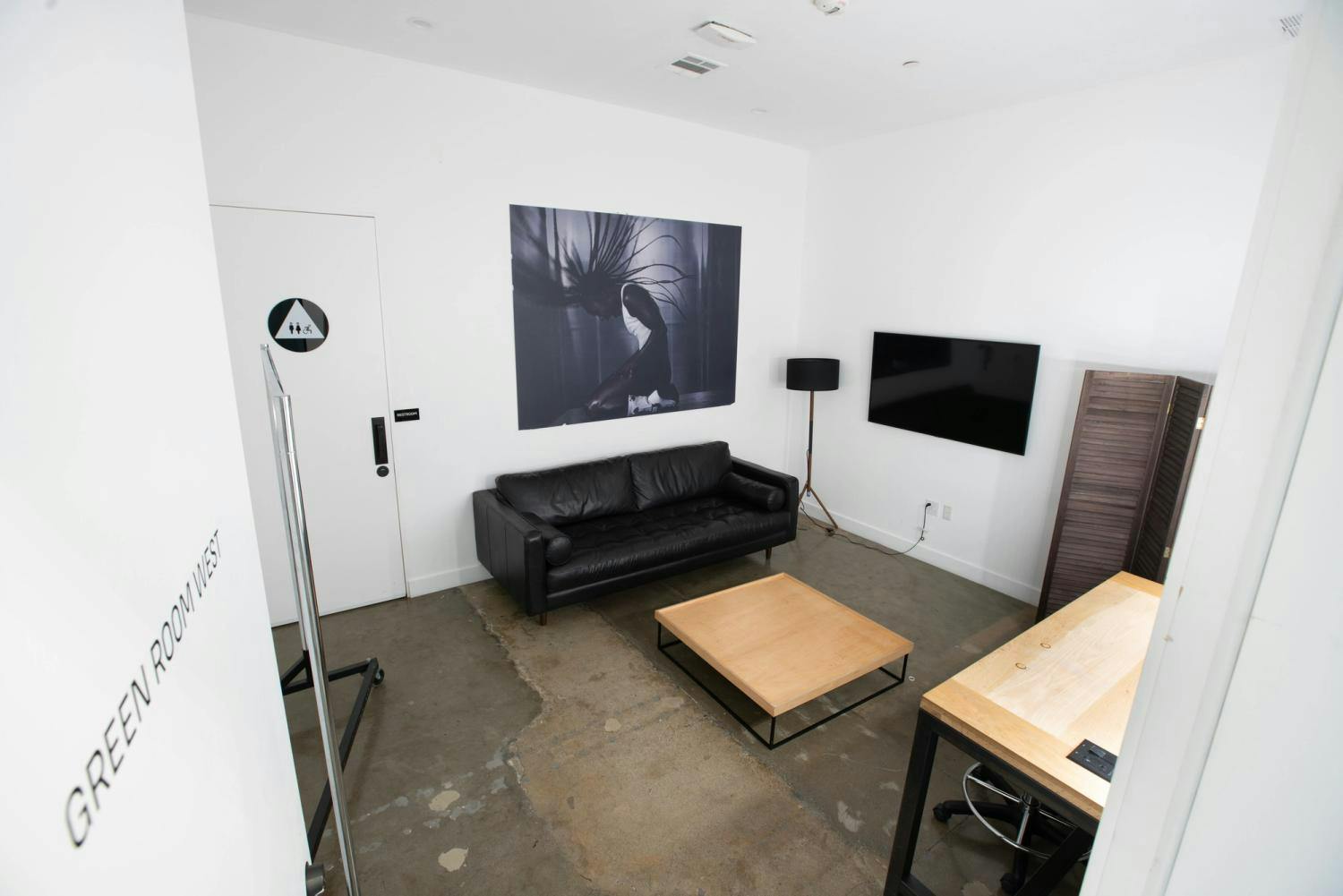 A clean, modern green room with a black leather couch, wooden coffee table, flat-screen TV, and a large canvas print, viewed from behind a glass partition with "GREEN ROOM WEST" lettering.