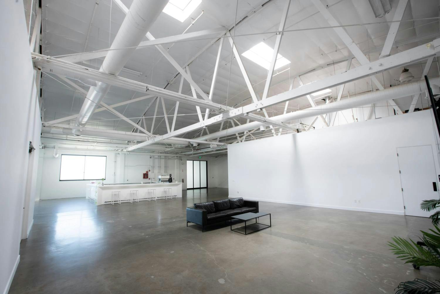 A large, airy gallery space featuring a black leather couch, white walls for artwork, and a natural light-filled environment with industrial accents.