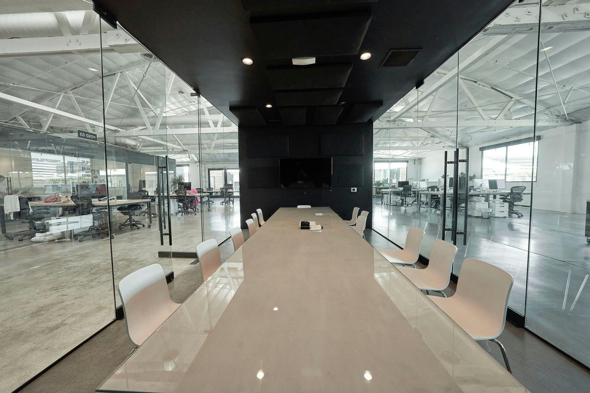 A modern conference room with a long table and chairs, glass walls, and a view into a workspace with desks and computers.