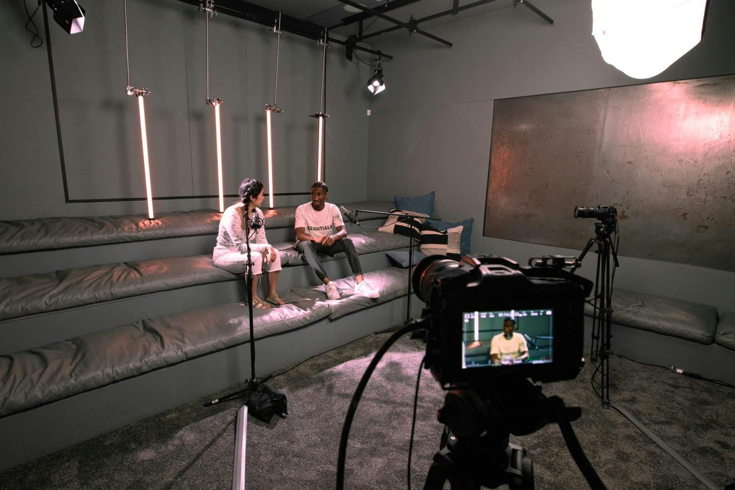 A wide-angle shot of a studio interview set with a male and female guest seated on a cushioned bench with vertical lights in the background.