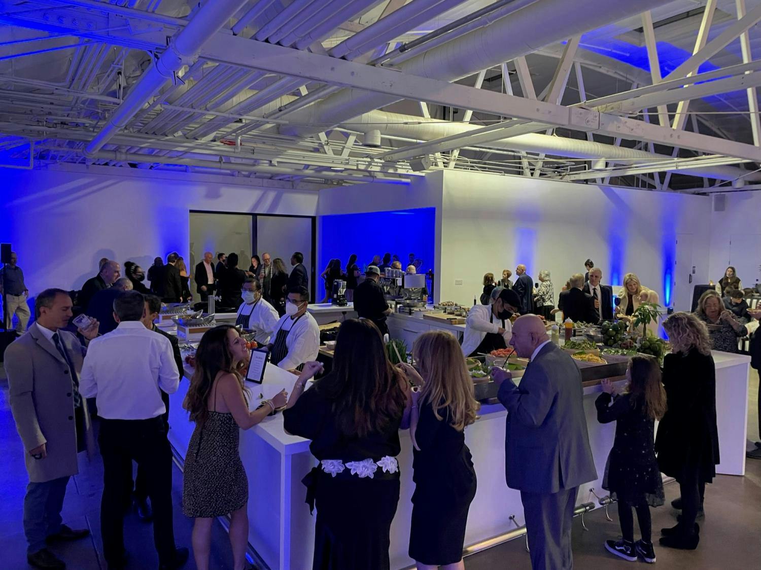 A bustling gallery event with guests mingling around a well-appointed buffet under soft blue lighting and industrial beams.
