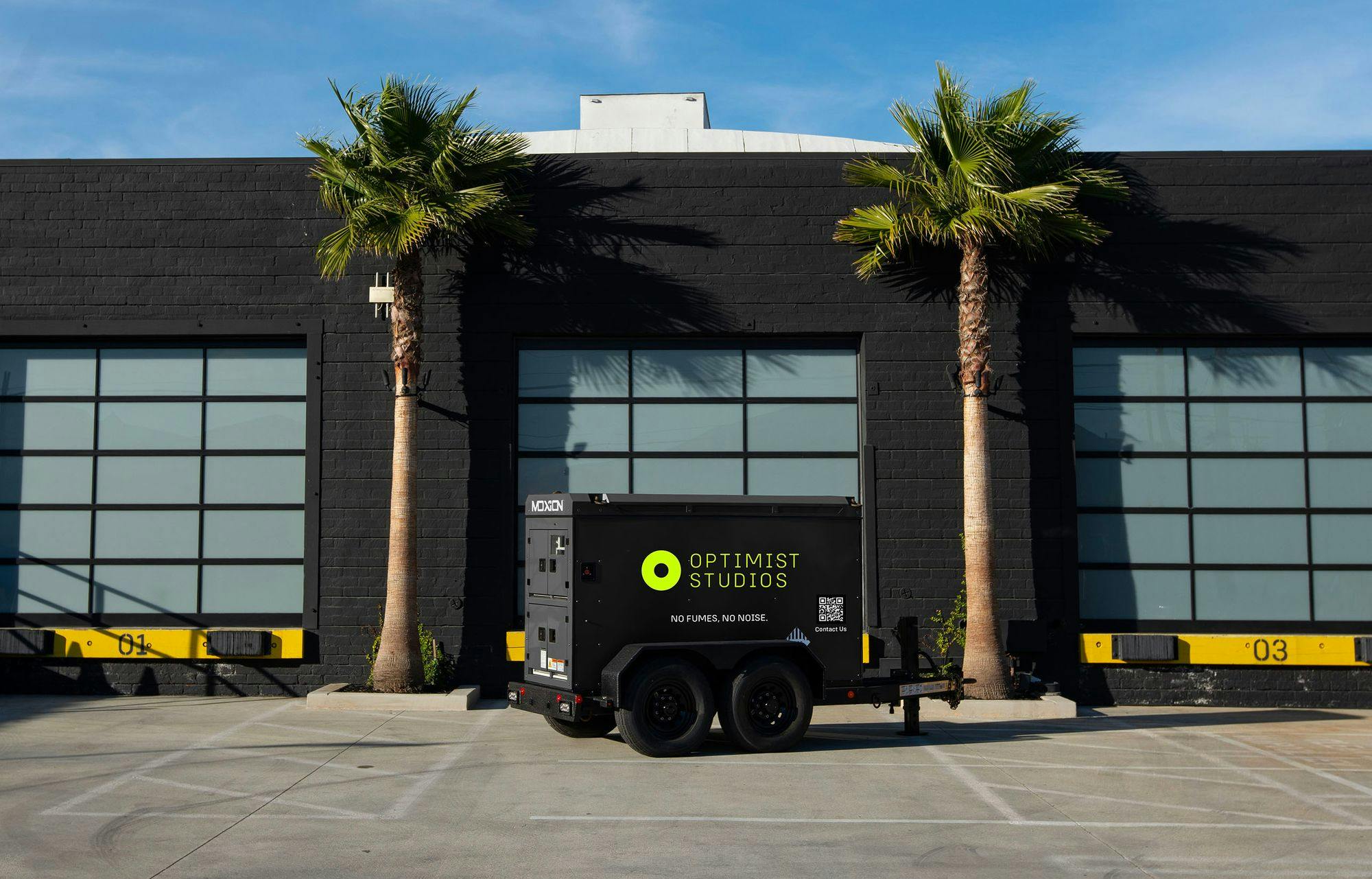 Image features a Moxion generator trailer parked in front of a building with a modern and eco-conscious design. The generator is marked with the logo and branding of "OPTIMIST STUDIOS," emphasizing their commitment to environmentally friendly practices with the slogans "NO FUMES, NO NOISE."