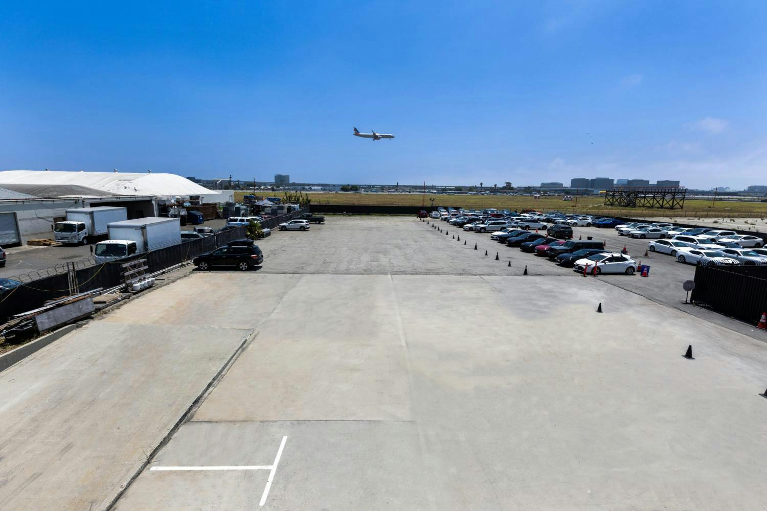 Wide-angle view of a large, open parking area filled with cars and trucks, with an airplane flying overhead near industrial buildings.