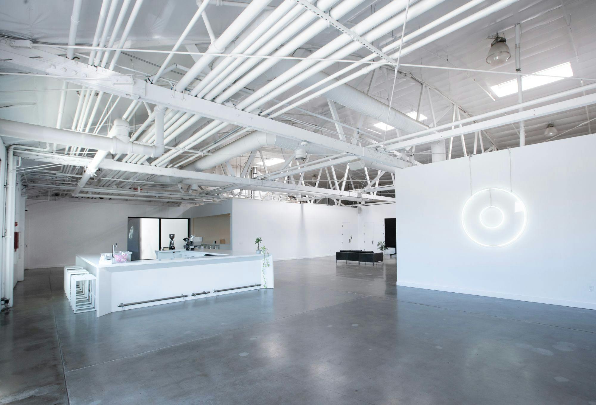 A spacious industrial-style gallery with white walls, high ceilings with exposed ductwork, and a minimalist bar area illuminated by a modern circular light fixture.