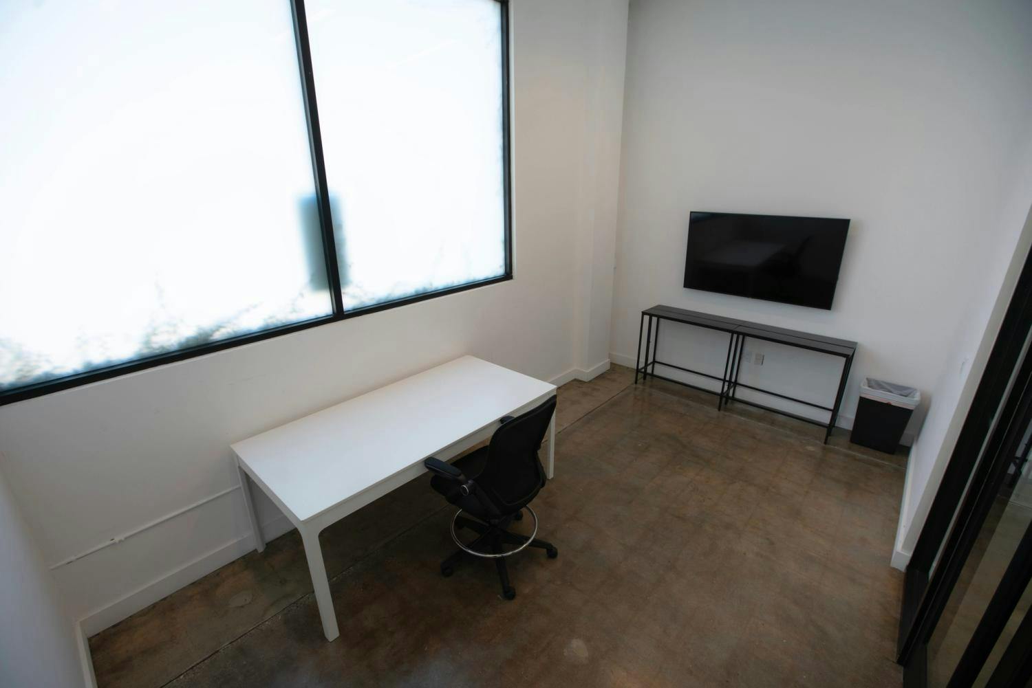 A minimalist workspace with a white desk and chair, a flat-screen TV on the wall, and a large window filling the room with natural light.