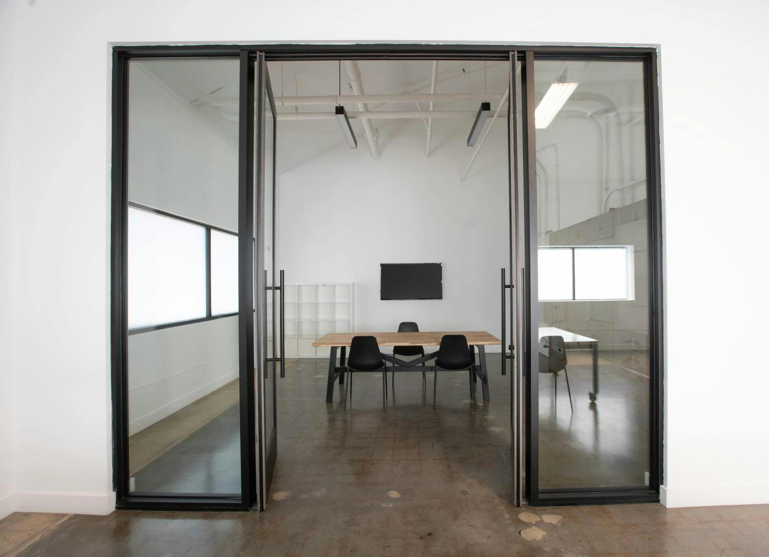 Through open glass doors, a modern office with a wooden table, black chairs, and a TV is framed by an industrial-style interior.