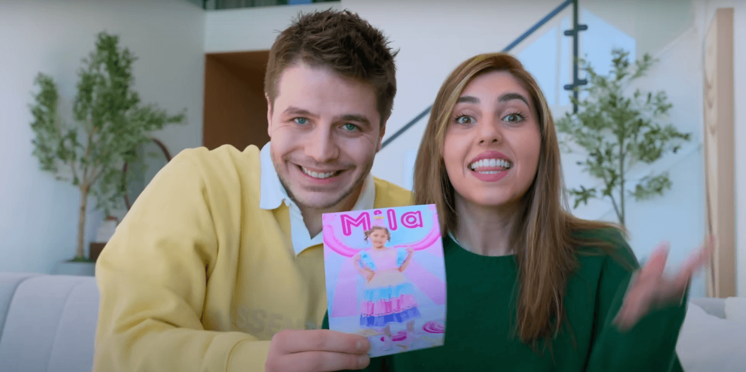 A smiling young couple, with the man holding a magazine featuring a child model, enthusiastically engage with the camera in a modern living room setting.