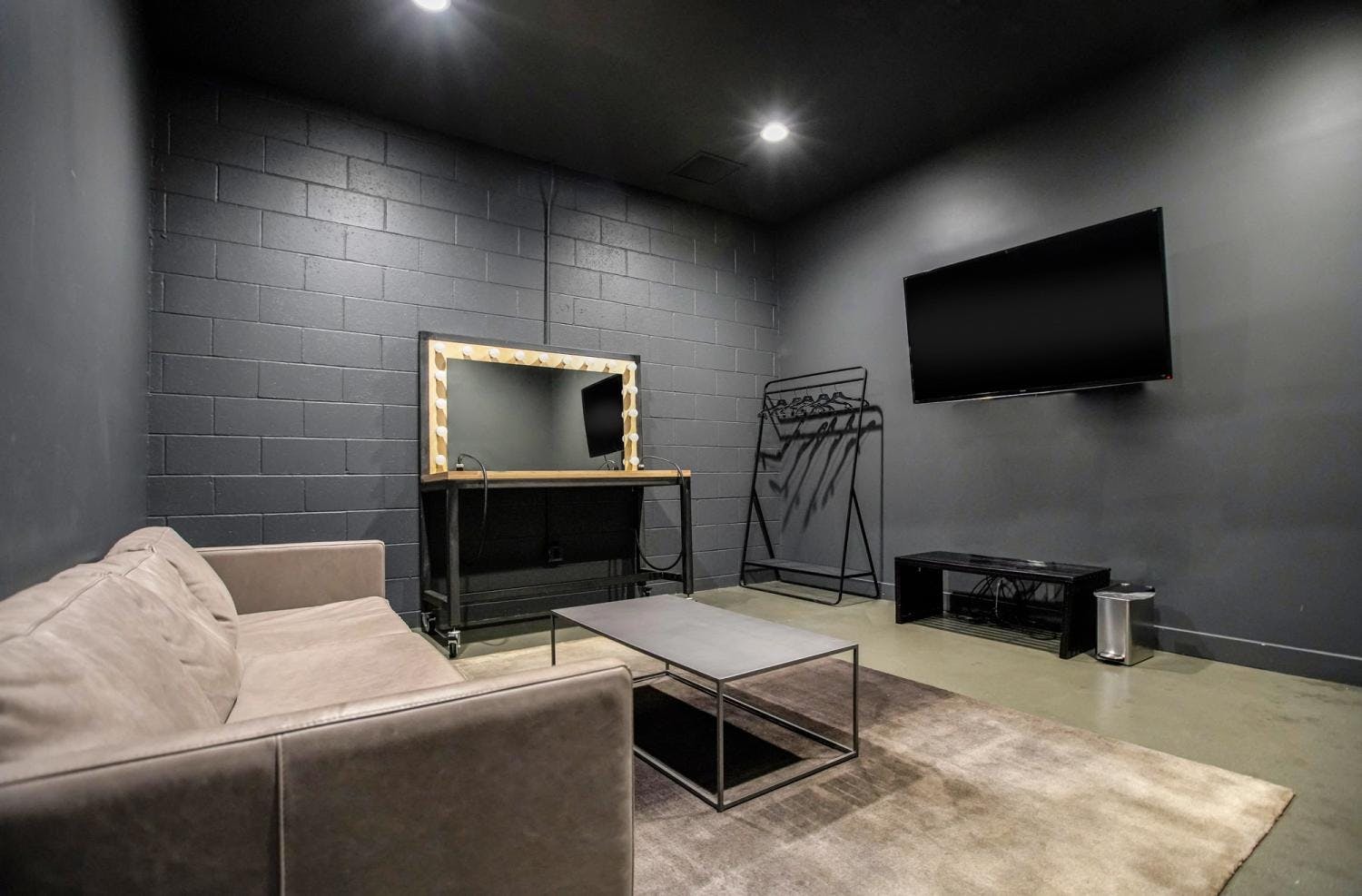 A dressing room with a beige couch, grey coffee table, makeup desk with illuminated mirror, and a TV.