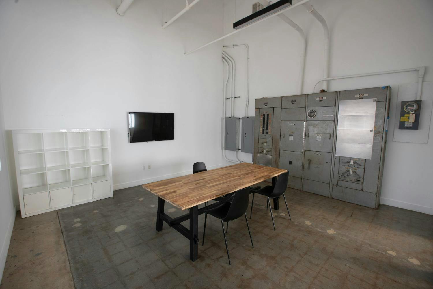  A functional workspace featuring a wooden table with black chairs, a wall-mounted TV, and a large metal utility cabinet, set against industrial flooring.