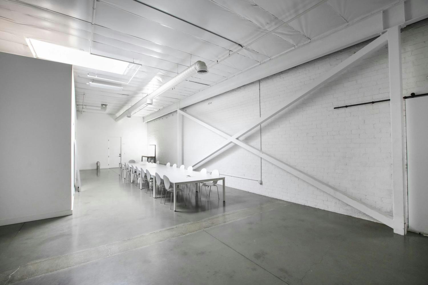 A bright, minimalist room featuring a meeting area with a long white table and chairs, white brick walls, and a polished concrete floor, illuminated by natural light and overhead fluorescent lights.