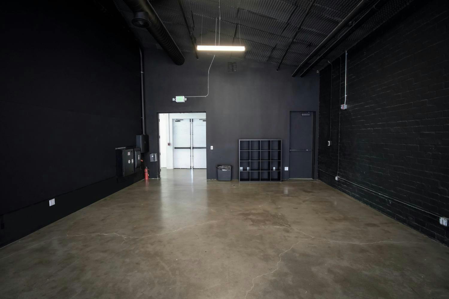 An empty corner of a studio showing black walls, concrete flooring, a storage cubby, and the entryway with industrial-style ceiling and lighting.