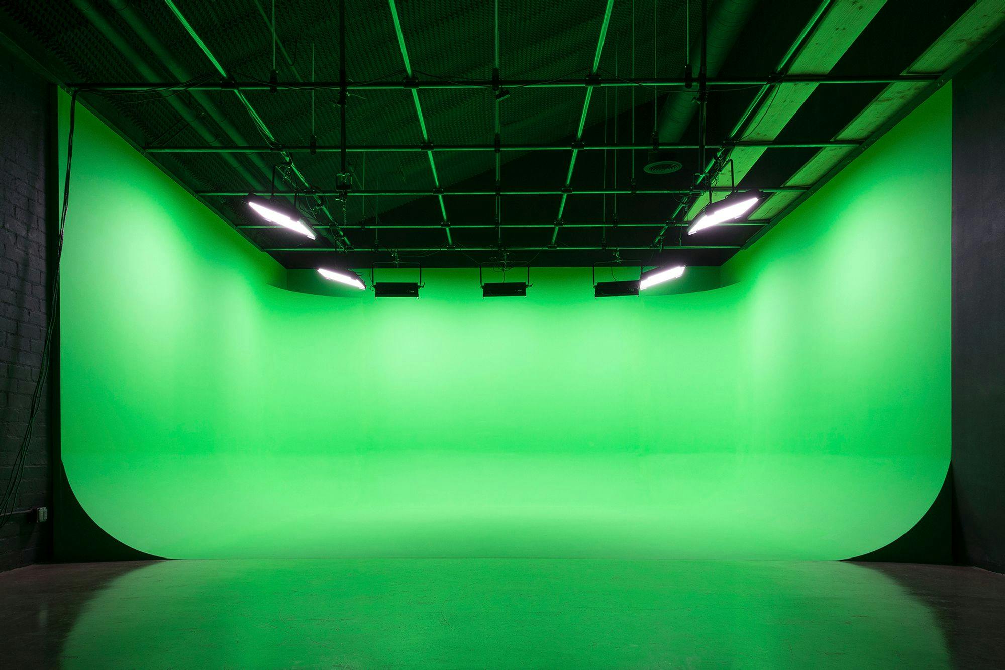 A modern studio with vibrant green cyclorama for chroma key effects, featuring overhead lighting fixtures on a dark ceiling and a concrete floor.