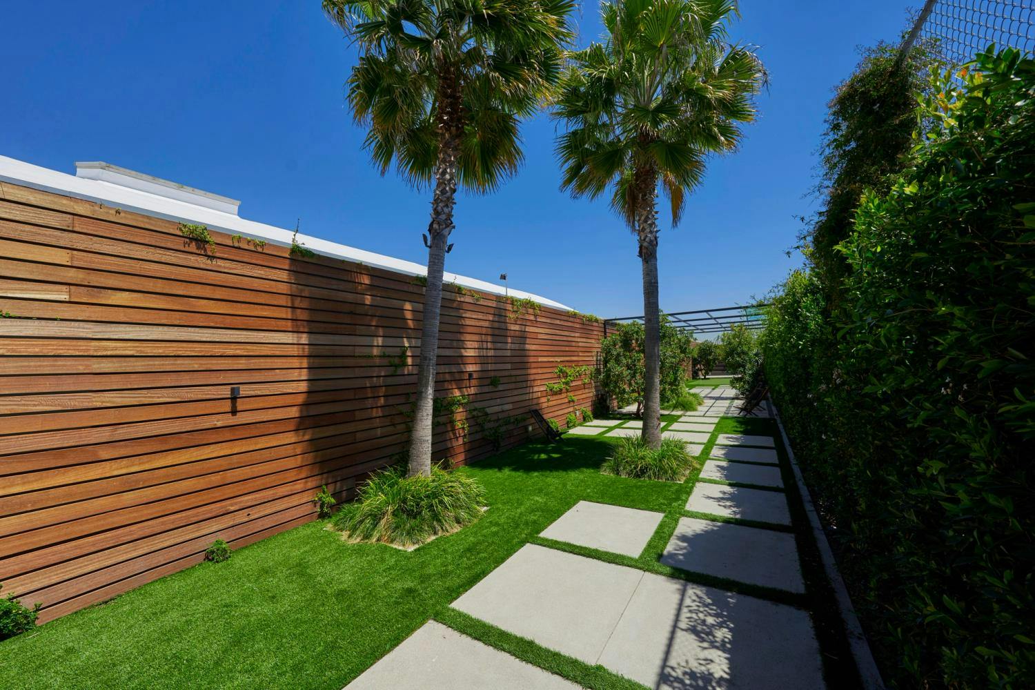 A serene garden pathway flanked by wooden fences and tall palm trees under a clear blue sky.